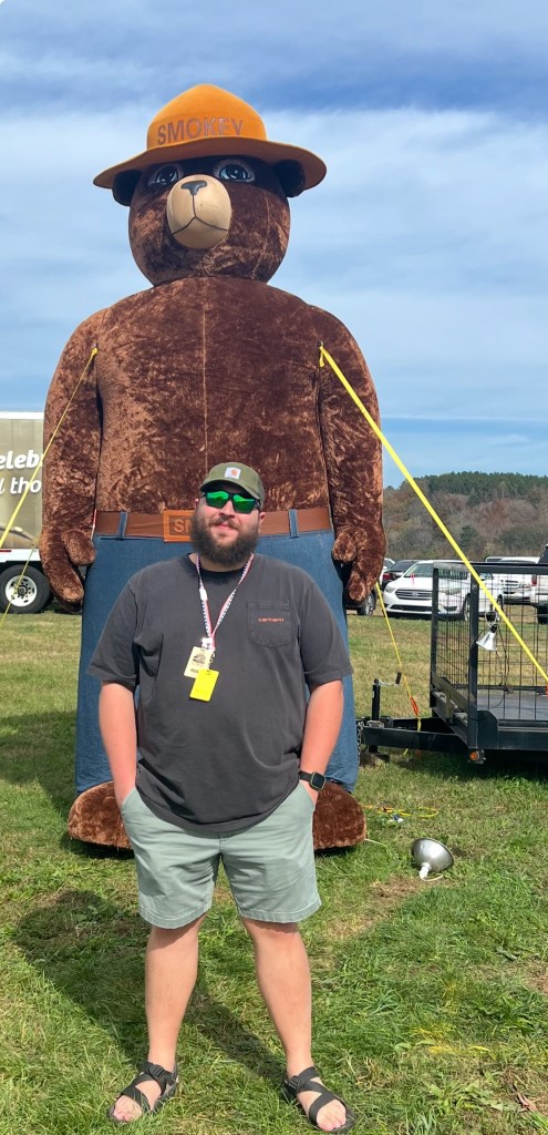 Nick Cruz standing outdoors on grass in front of a large Smokey Bear display, wearing sunglasses, a cap, and casual clothing at what appears to be an outdoor event.